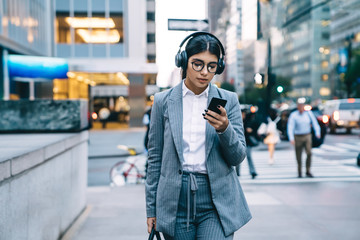 Thoughtful female in headphones browsing smartphone on street