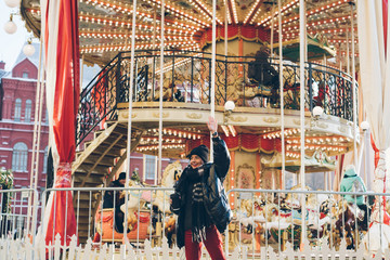 Cheerful young woman standing by illuminated carousel and waving with hand