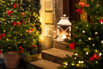 Fototapeta na wymiar Christmas trees decorated with garland lightings and bowknots of red ribbon, vintage lantern outdoors on doorsteps in european old town house on christmas eve