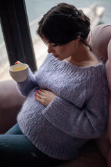 Young pregnant woman sit in cozy cafe with beautiful view of window of the sea and mountains drink healthy tea . Female dressed warm woolen sweater and siting on the sofa.