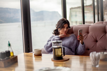 Young pregnant woman sit in cozy cafe with beautiful view of window of the sea and mountains drink healthy tea . Female dressed warm woolen sweater and siting on the sofa and writing massege on phone.