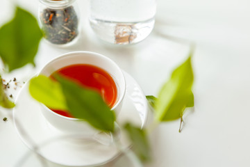 Cup of hot black tea on white table with dry tea jar