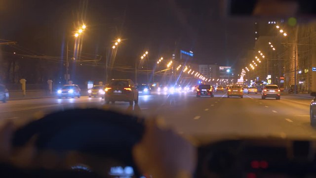 Night city street through the windshield of the moving car. Close up man's hands on the steering wheel of a car. POV.