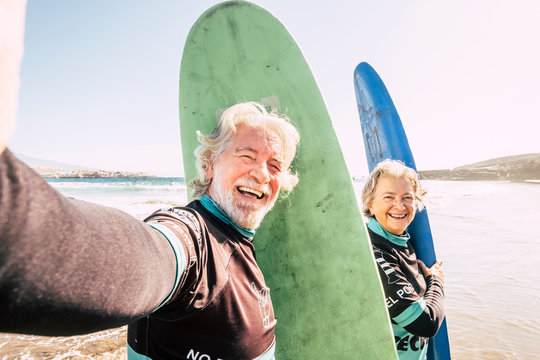 happy couple of seniors at the beach trying to go surf and having fun together - mature woman and man married taking a selfie with the wetsuits and surftables with sea or ocean at the background