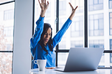 Energetic ethnic businesswoman celebrating with hands up at office table