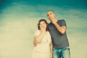 A plump red-haired attractive woman and a tall bald man on a blue background are emotionally surprised at what they saw. They look at the camera and closed their mouth with  hands.
