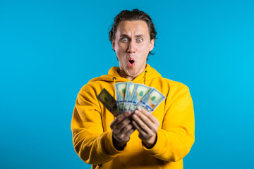 Man in yellow hoodie with surprised happy face holding US currency. Person with money. New dollars in hands on blue studio background .
