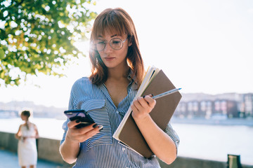 Young redhead woman in eyewear for vision protection read income messages on smartphone strolling in city, charming millennial caucasian female student check email and notification on cellphone.
