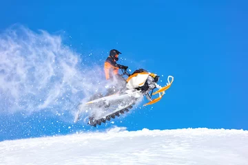Fotobehang elite sports snowmobiler rides and jumps on steep mountain slope with swirls of snow storm. background of blue sky leaving a trail of splashes of white snow. bright snowmobile and suit without brands © Wlad Go