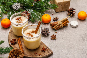 Eggnog with cinnamon and nutmeg for Christmas and winter holidays. Homemade beverage in glasses with spicy rim. Tangerines, candles, gift. Stone concrete background