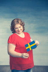 Cute smiling chubby red-haired woman holding a swedish flag on a blue background