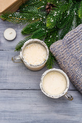 Eggnog cocktail in two glasses mug arranged with christmas decoration on light wooden table. Evergreen fir tree branch, gift, cozy jersey, artificial snow