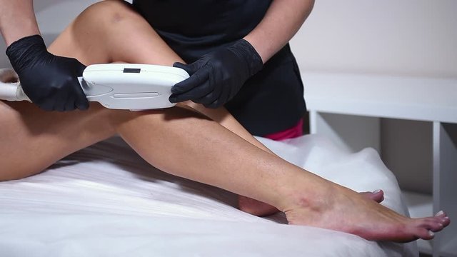 Laser hair removal on woman legs. The girl is lying on the couch in the medical glasses in the treatment room. The hands of the cosmetologist or beautician making epilation of the legs