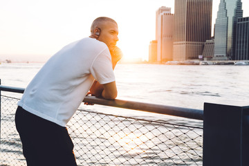 Young ethnic man with earphones leaning on handrail against city
