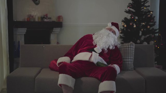 Portrait of mature Caucasian man in Santa Claus costume sleeping on the couch holding the bottle of beer. Old guy spending holidays alone at home. Bad santa, loneliness, depression, alcohol addiction.
