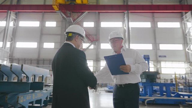 Businessmen greet each other shake hands on background of modern factory facility. Multi racial engineers contractors in helmets meet with handshake at workshop