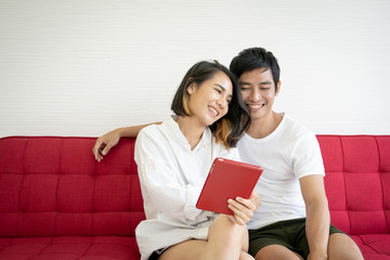 Fototapeta premium Asian young couple sitting on the red sofa in living room and using digital tablet close up.