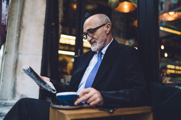 Senior bearded businessman in glasses with coffee cup reading newspaper