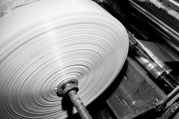 Black & White imagery of spool of plastic packaging sheet heavy duty for industrial use outdoors. wrapped around a metal pole ready to feed into a machine for cutting into shape or printing logos.