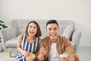 Portrait of a laughing young Asian couple hugging while sitting together at home and watching TV
