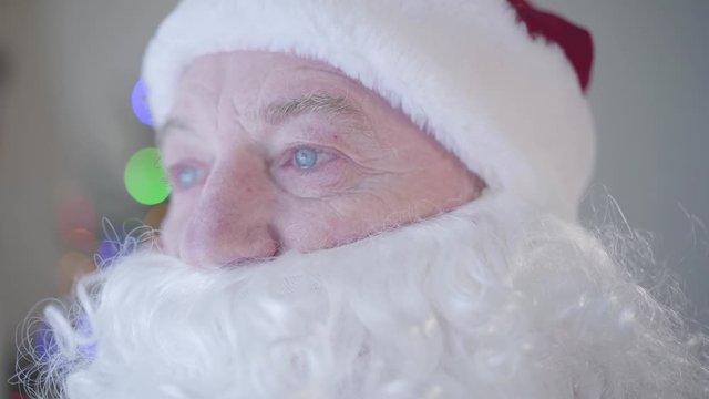 Close-up portrait of the old man with false beard in costume of Santa Claus crying standing in front of New Year tree. Tears appear on the man's eyes. Concept of sadness, sadness, lack of festive mood
