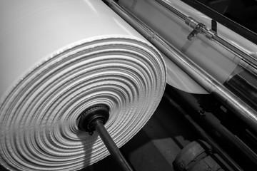 Black & White imagery of spool of plastic packaging sheet heavy duty for industrial use outdoors. wrapped around a metal pole ready to feed into a machine for cutting into shape or printing logos.