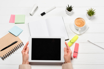 cropped view of woman holding digital tablet at workplace with office supplies and coffee