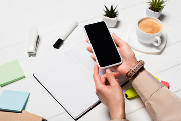 cropped view of woman holding smartphone at workplace with office supplies and coffee