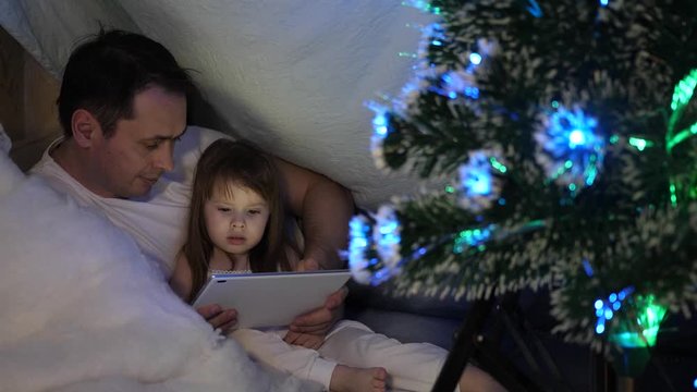 Father and daughter on Christmas evening, play and watch cartoons on tablet, in a children's room in tent with garlands. baby and dad are playing in room. concept of happy childhood and family.