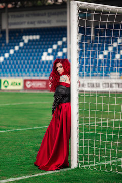 Luxurious woman with red hair and in a red dress plays on the football field.
