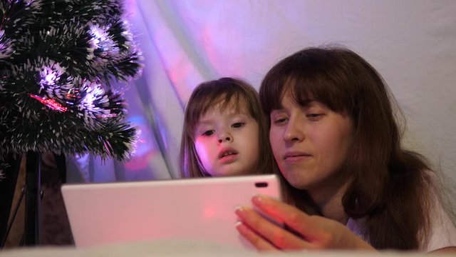 mom and daughter on Christmas evening, play and watch cartoons on tablet, in a children's room in tent with garlands. baby and mother are playing in the room. concept of happy childhood and family.