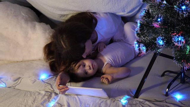 happy mother and daughter on Christmas evening, play cartoons and watch cartoons on tablet, in children's room in a tent with garlands. baby and mother are playing in room. happy childhood concept.