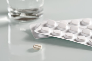 White still life with an antibiotic pill lying on a glass plate in front of two blister packs with tablets and a glass of water