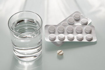 White still life with two blister packs with antibiotic tablets and a single pill lying on the background together with a glass of water