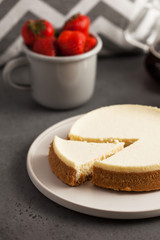 Coconut cheesecake on a round plate. In the background, a teapot with black coffee and a mug of fresh strawberries. Food photo on gray background.