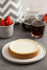 Coconut cheesecake on a round plate. In the background, a teapot with black coffee and a mug of fresh strawberries. Food photo on gray background.
