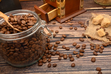 Wooden spoon in a glass jar with coffee beans, coffee grinder, pieces of brown sugar on a wooden background. Close up.