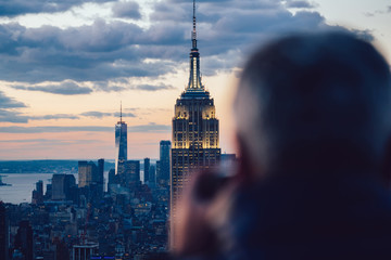 Obraz premium Back view of male traveler taking photo of skyscrapers in skyline at evening time exploring New York city during holidays. Sightseeing to Famous Empire State Building Landmark. Scene cityscape view