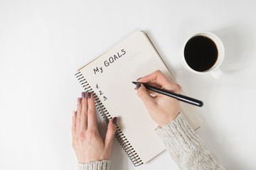 Female hands writing My Goals in a notebook. Mug of coffee on the table, top view