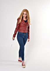 Full length portrait of a pretty blonde girl wearing red leather jacket denim jeans and sneakers. Standing pose, holding a dagger,  on a studio background.