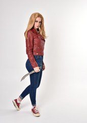 Full length portrait of a pretty blonde girl wearing red leather jacket denim jeans and sneakers. Standing pose, holding a dagger,  on a studio background.