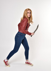Fototapeta na wymiar Full length portrait of a pretty blonde girl wearing red leather jacket denim jeans and sneakers. Standing pose, holding a dagger, on a studio background.