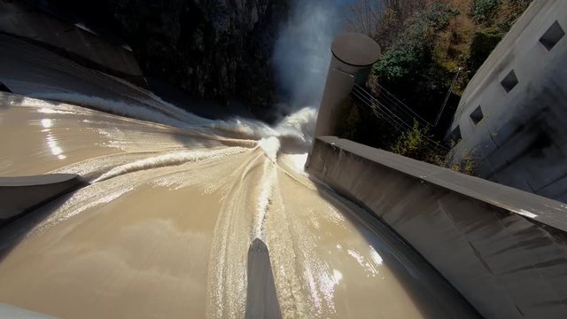 Slow motion left truck moving looking down dirty brown yellow flooded river water flowing over concrete spillway