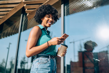 Cheerful african american woman with curly hair holding mobile phone sending text messages chatting...