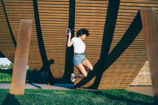 Overjoyed african american millennial woman jumping excited with sound of favorite songs in earphones, happy dark  skinned hipster girl entertaining with player jumping up feel carefree in park