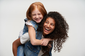 Lovely red haired little girl sitting on the back of her cheerful dark skinned curly female friend and smiling happily, having fun together while posing over white background