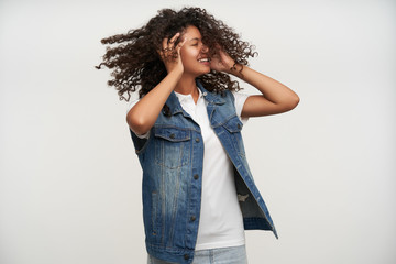 Pleasant looking young dark skinned long haired curly woman turning her head and keeping eyes closed, smiling happily while posing over white background with raised hands