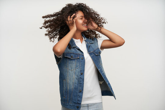 Pleasant looking young dark skinned long haired curly woman turning her head and keeping eyes closed, smiling happily while posing over white background with raised hands