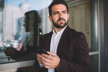 Pensive caucasian man in formal outfit holding smartphone pondering on smth planning to send message, serious male in trendy jacket looking away using mobile phone and 4G for browsing outdoors.