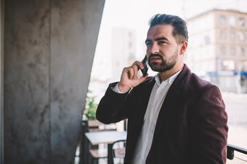 Pensive caucasian man entrepreneur talking on mobile phone standing outdoors, prosperous confident male in trendy jacket making smartphone call using 4G concentrated on business conversation